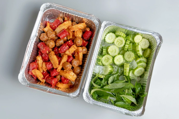 Two foil containers with a prepared meal and fresh vegetables.